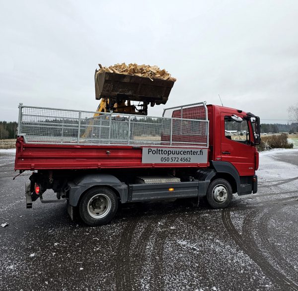 Traktorin kauha laittamassa polttopuita pienen punaisen kuorma-auton lavalle.